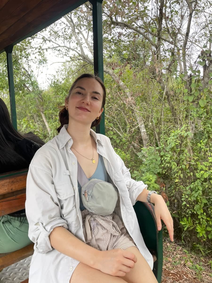 Allie, seated under a wooden canopy, with green trees behind